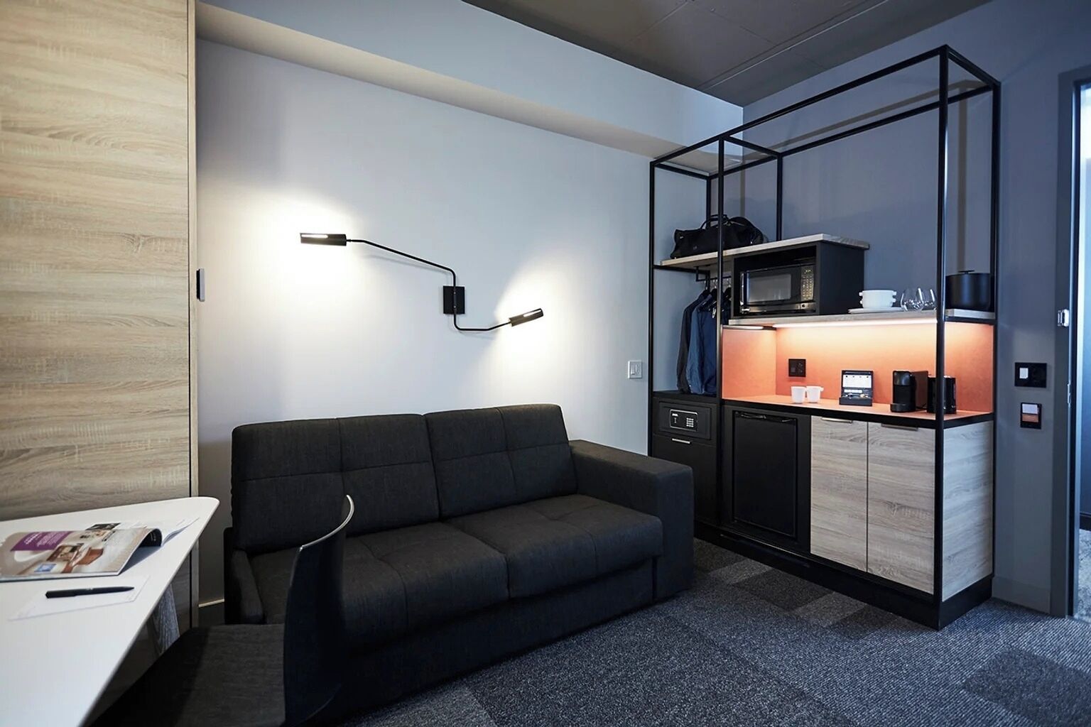 Habitació d'hotel senzilla amb sofà, escriptori petit, zona de cuina i separador de vidre.
