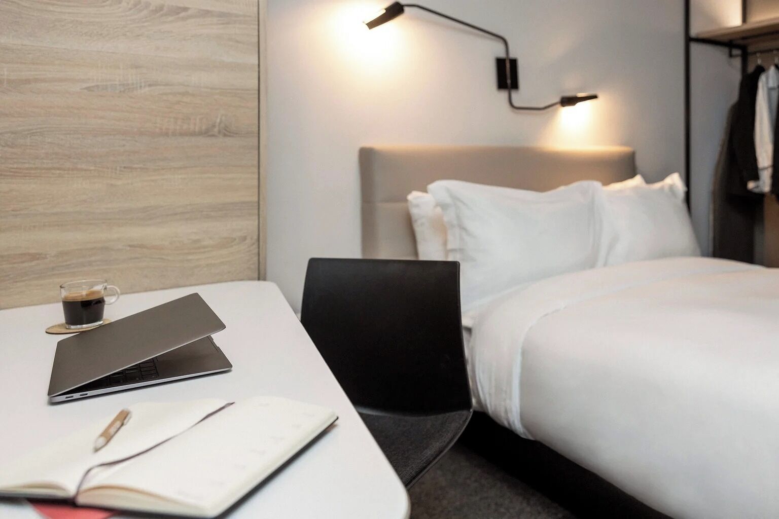 Una habitación de hotel con una cama, un escritorio con una computadora portátil, una taza de café y un cuaderno.