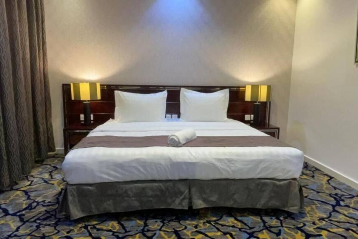 A hotel room with a large bed, a headboard with lights, a patterned carpet, and a painting on the wall.