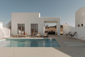 Un área de piscina moderna al aire libre con piscina azul cristalina, sillas de descanso y una zona de estar con mesa y sillas.