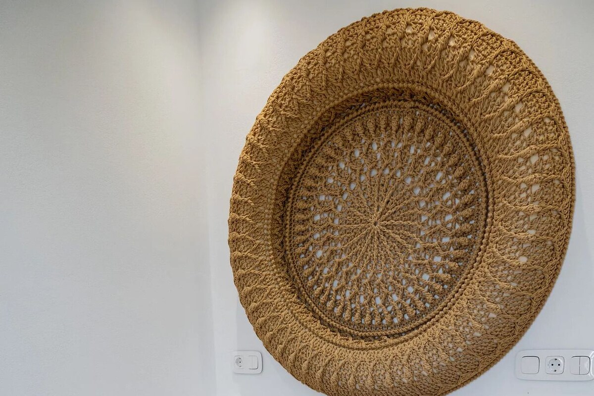 Una cama con colcha estampada, dos almohadas, un tapiz de pared tejido y un sombrero de paja sobre la cama.