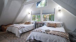 A cozy attic bedroom with two beds, a desk, and large windows offering a forest view.