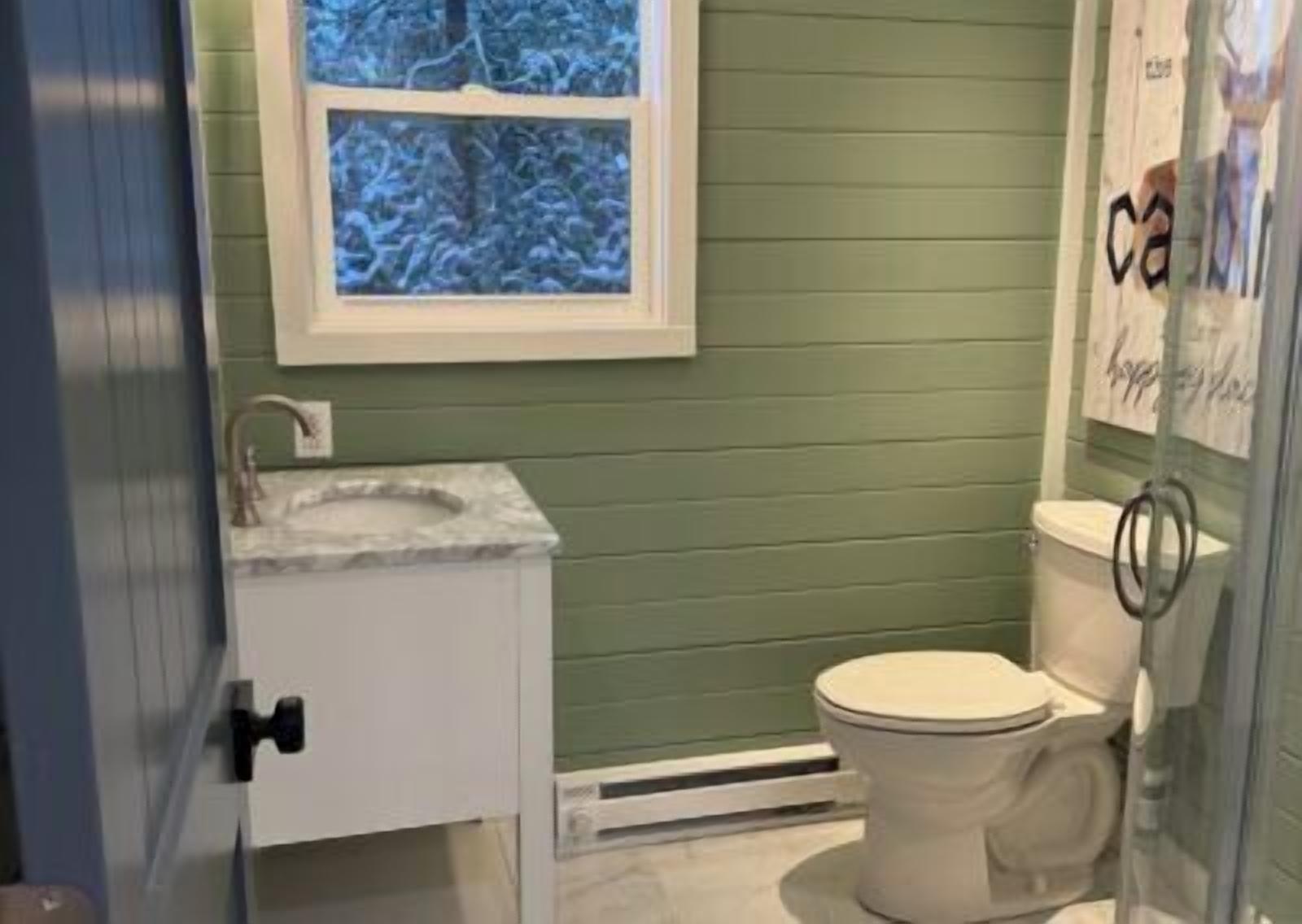Et badeværelse med grøn væg, hvid toilet, marmorvask og et vindue med udsigt til træer.