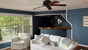A room with a white sofa, a white armchair, a ceiling fan, and a window with a view of trees.