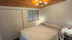 Un dormitorio con techo de madera, una cama con colcha estampada, una mesita de noche con lámpara y una ventana con persianas.