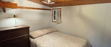 Quarto com teto de madeira, uma cama com colcha bege, mesa de cabeceira com abajur e cômoda.