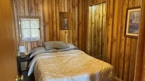 Una habitación con paneles de madera, una cama con cubrecama blanco, una mesita de noche con lámpara y una ventana con cortinas.
