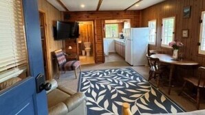 Interior de cabaña con detalles de madera, un televisor montado en la pared, zona de comedor con mesa y sillas, y una alfombra estampada.
