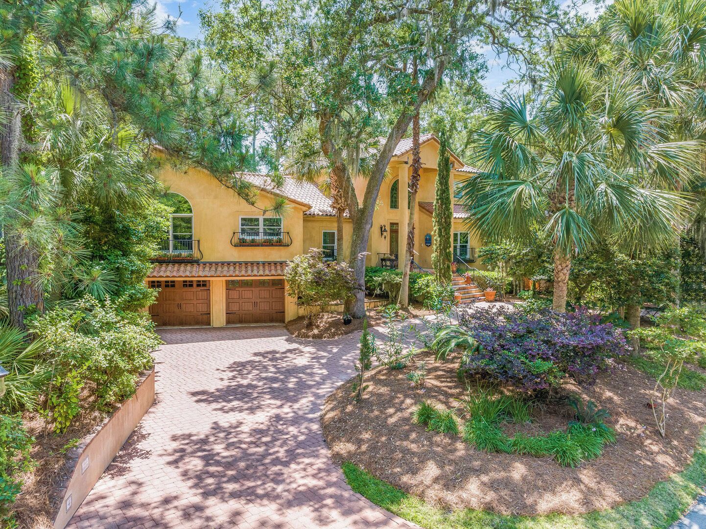 A yellow house with a brick driveway, surrounded by lush greenery and palm trees.
