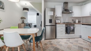 Eine moderne Küche mit weißen Schränken, einem Edelstahlkühlschrank und einem eingebauten Backofen. Es gibt eine Essecke mit einem weißen Tisch und Stühlen.