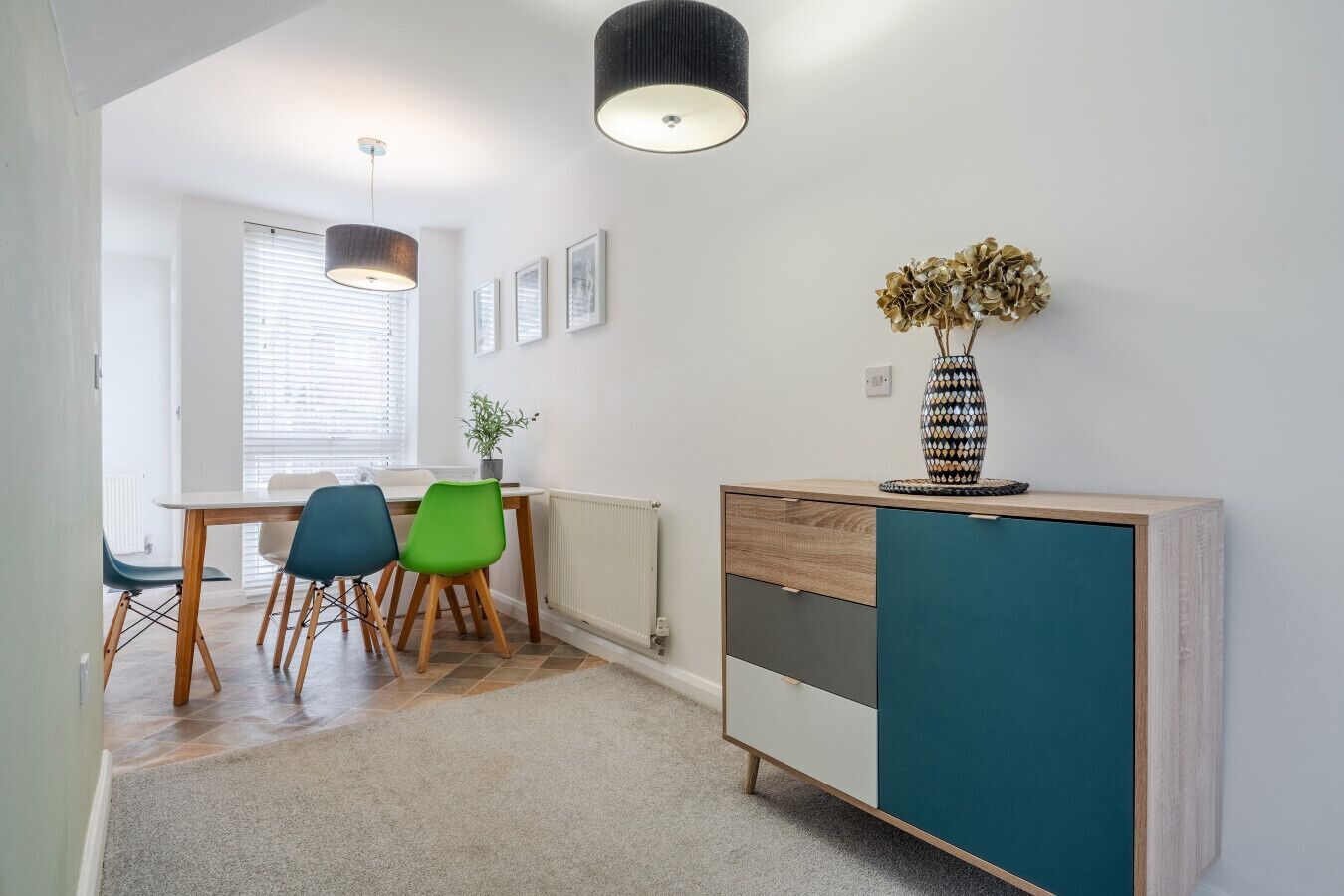 A modern kitchen with a dining area, featuring a wooden table, two chairs, a cabinet, and a vase with dried flowers.