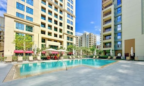 A swimming pool surrounded by modern apartment buildings, outdoor seating, and umbrellas.