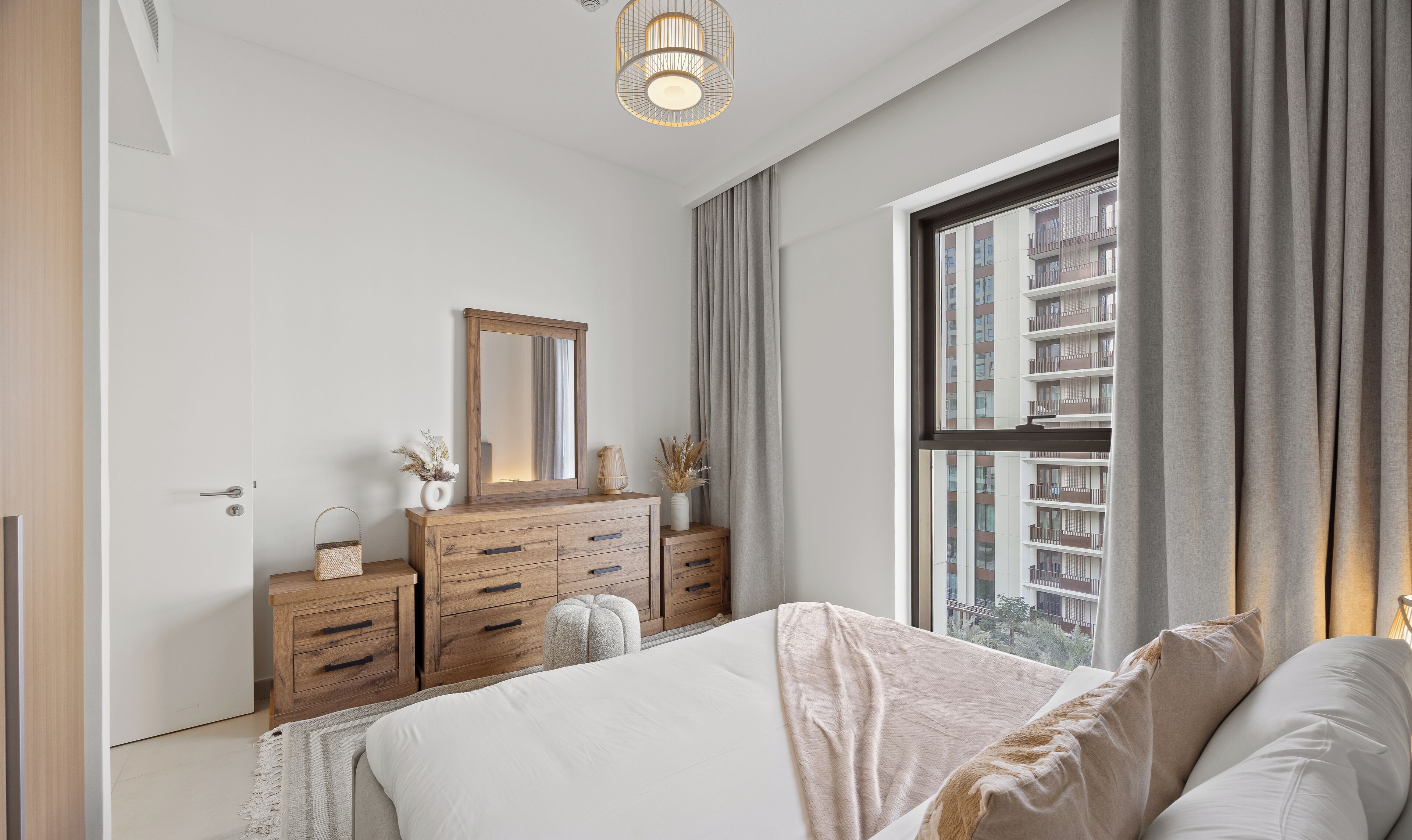 A bedroom with a bed, a dresser, a mirror, and a window with a view of buildings.