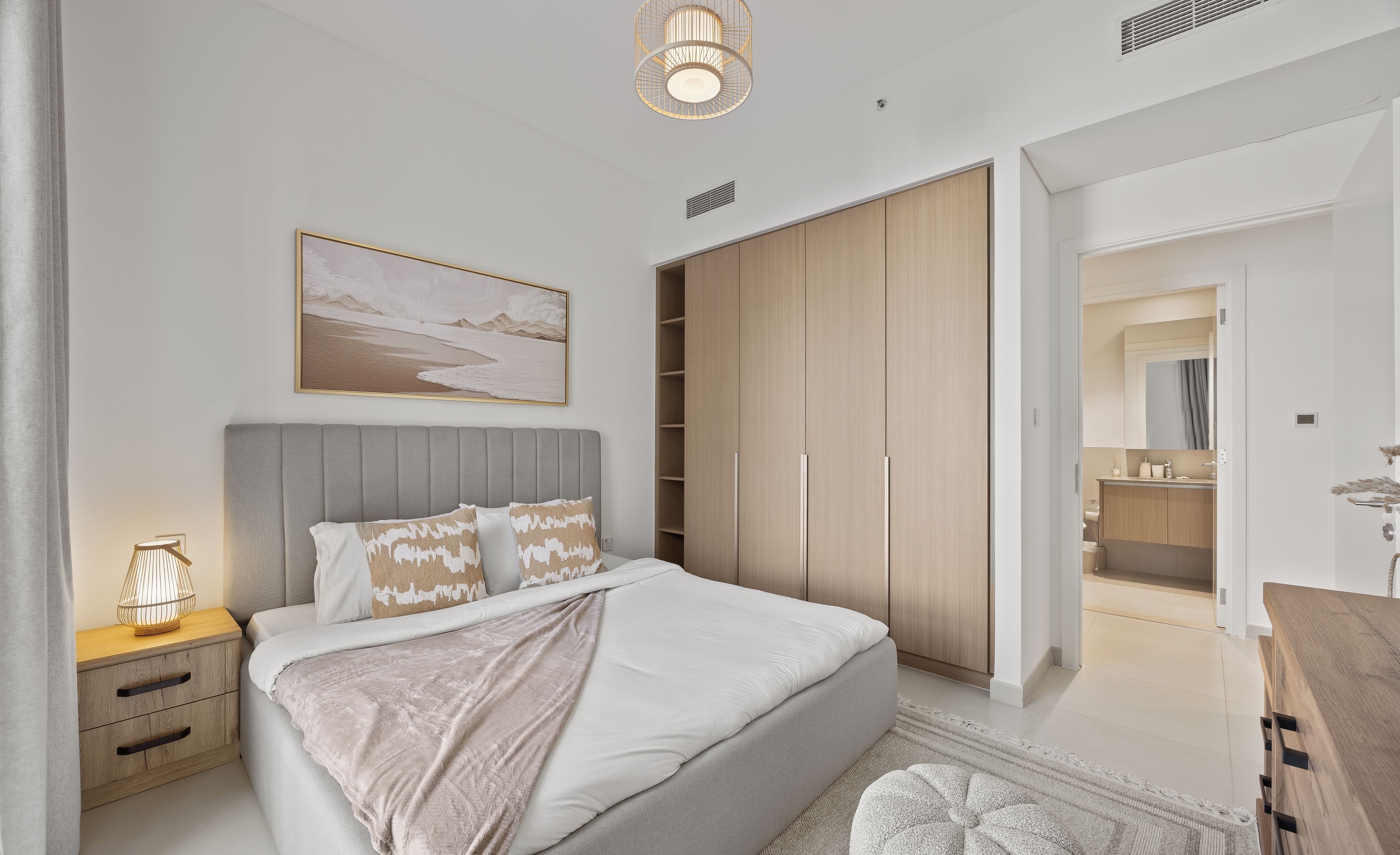 A modern bedroom with a large bed, a bedside table with a lamp, a wardrobe, and a bathroom visible through an open door.