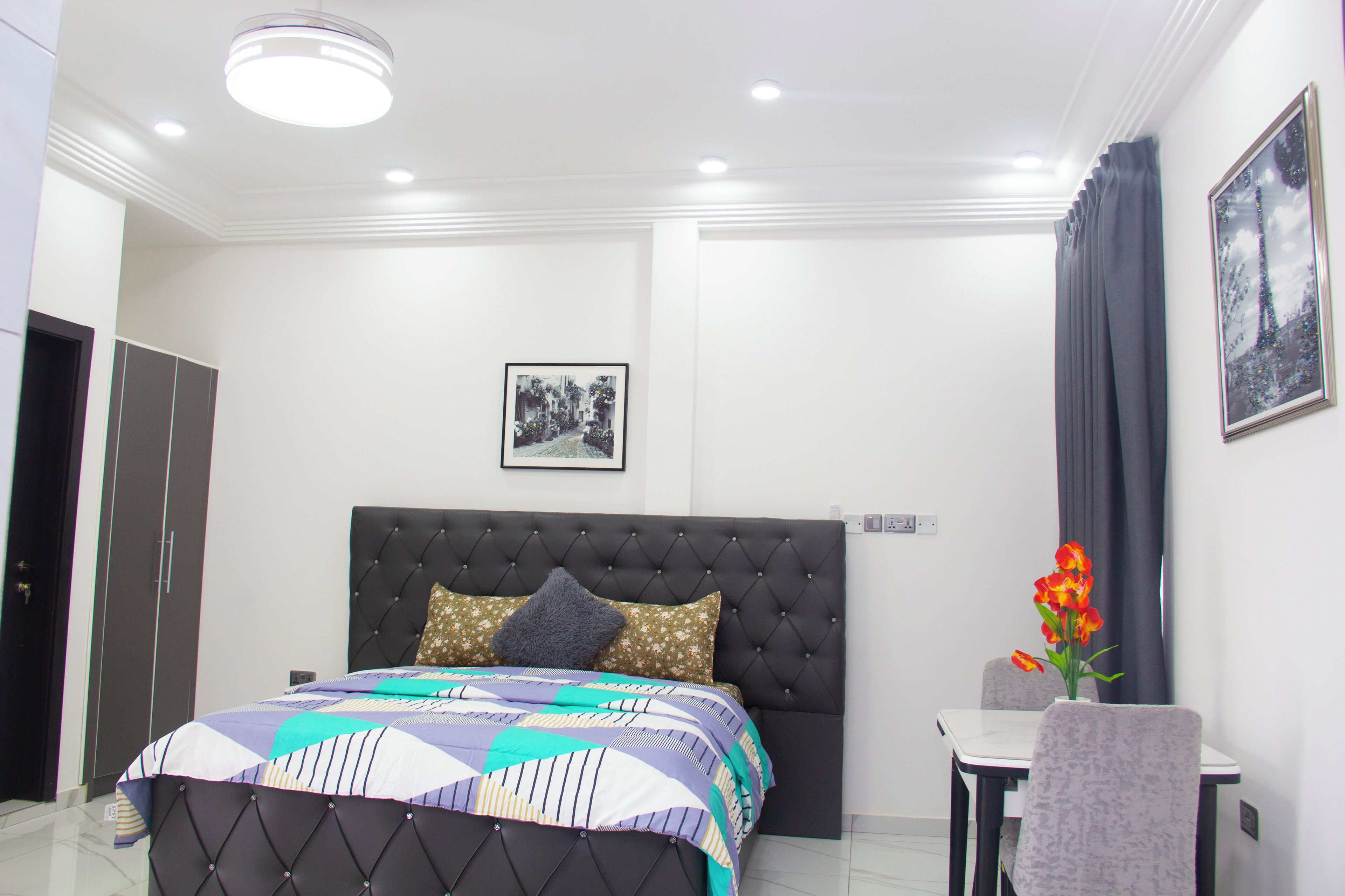 Un dormitorio con cama, mesita de noche, una silla, un cuadro en la pared y una ventana con cortinas.