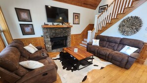 A living room with a stone fireplace, two brown sofas, a wooden coffee table, and a staircase with wooden railings.