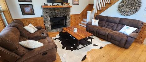 A living room with a stone fireplace, two brown sofas, a wooden coffee table, and a staircase with wooden railings.