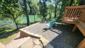 A stone patio with a fire pit, two blue chairs, and a wooden deck overlooking a lake.