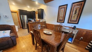 A spacious living room with a wooden dining table and chairs, a sofa, and a kitchen area with dark wooden cabinets and a refrigerator.