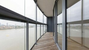 A corridor with large windows, wooden flooring, and a view of a river and cityscape.