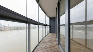 A corridor with large windows, wooden flooring, and a view of a river and cityscape.