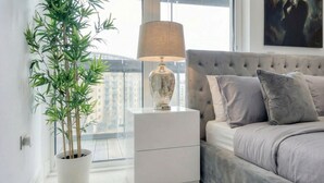 A bedroom with a bed, a nightstand with a lamp, a potted plant, and a city view.