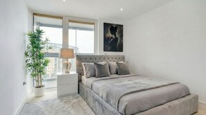 A bedroom with a large bed, a potted plant, a nightstand, a lamp, and a painting on the wall.