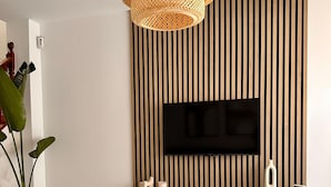 A modern living room with a flat-screen TV mounted on a wooden slat wall, a white console table, a woven pendant light, and a potted plant.