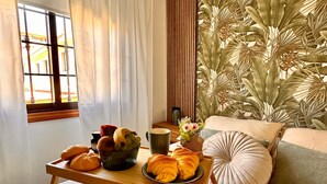 A cozy room with a wooden table set with pastries and a fruit bowl, a sofa with cushions, and a wall with a tropical leaf pattern.