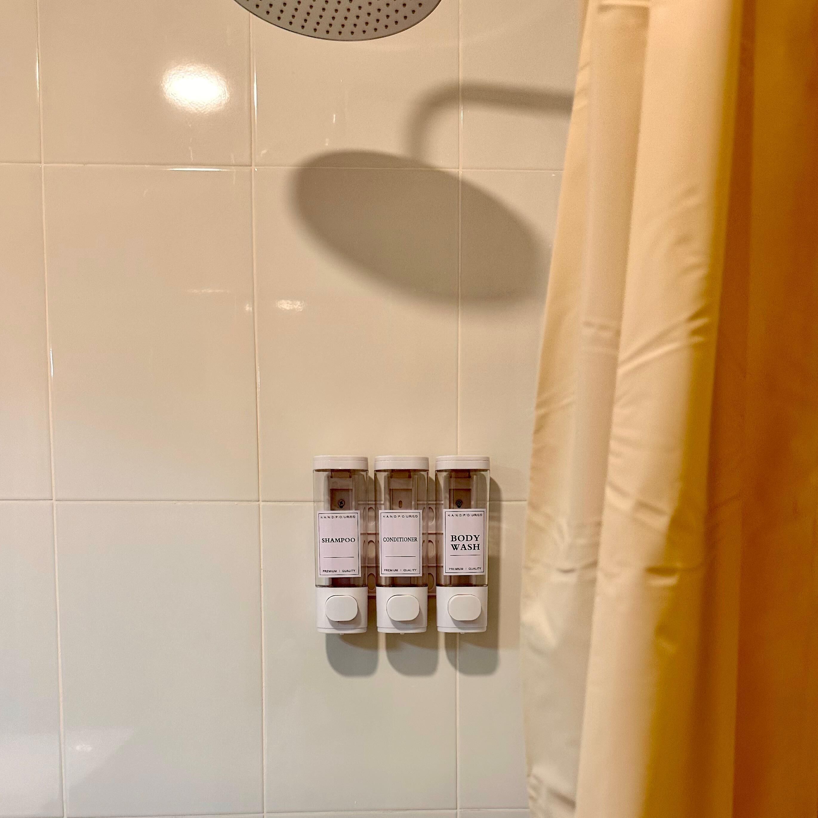 Een badkamer met een betegelde muur, een douchegordijn en drie dispensers met de aanduiding 'SHAMPOO', 'CONDITIONER' en 'BODY WASH'.