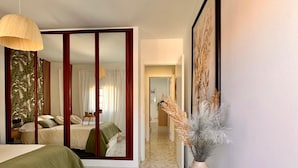A hotel room with a bed, a mirror, a vase with dried plants, and a hallway with a tiled floor.