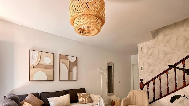 A modern living room with a dark brown sofa, a white armchair, and a wooden coffee table. There are abstract wall art pieces and a staircase with a wooden railing.