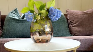 Une table ronde blanche avec un pied doré, un vase rempli de fleurs bleues et un livre posé sur l’étagère inférieure.