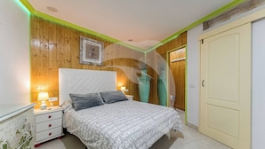 A bedroom with a bed, bedside tables, a nightstand, a lamp, a framed picture on the wall, and a door.