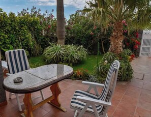 An outdoor patio with a round table and two striped chairs, surrounded by lush greenery and a palm tree. - Relax in El Médano's Modern Sea View Apartment (Granadilla de Abona)