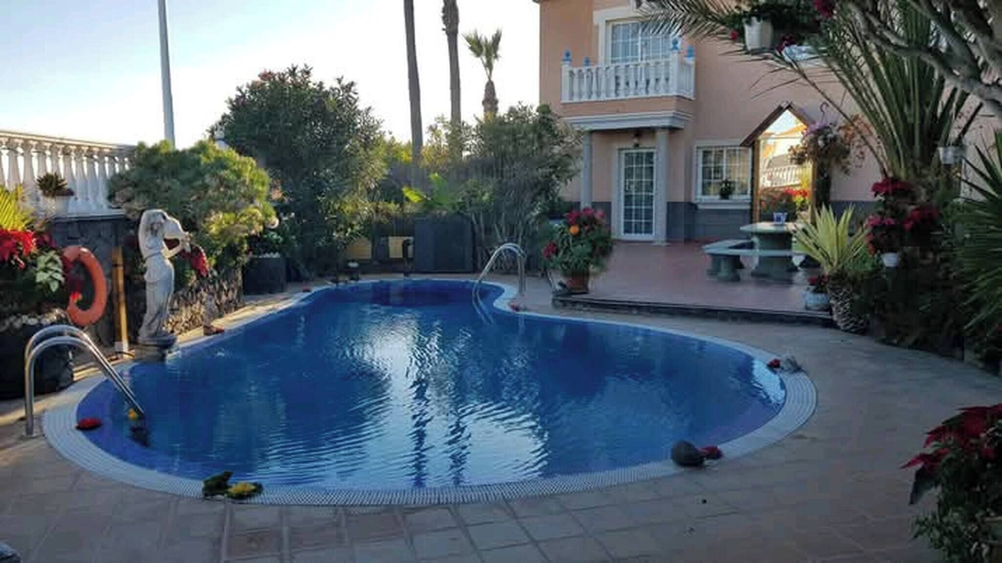 Ein Pool mit geschwungener Kante, umgeben von einer gepflasterten Terrasse, Palmen und einem Gebäude mit Balkon.