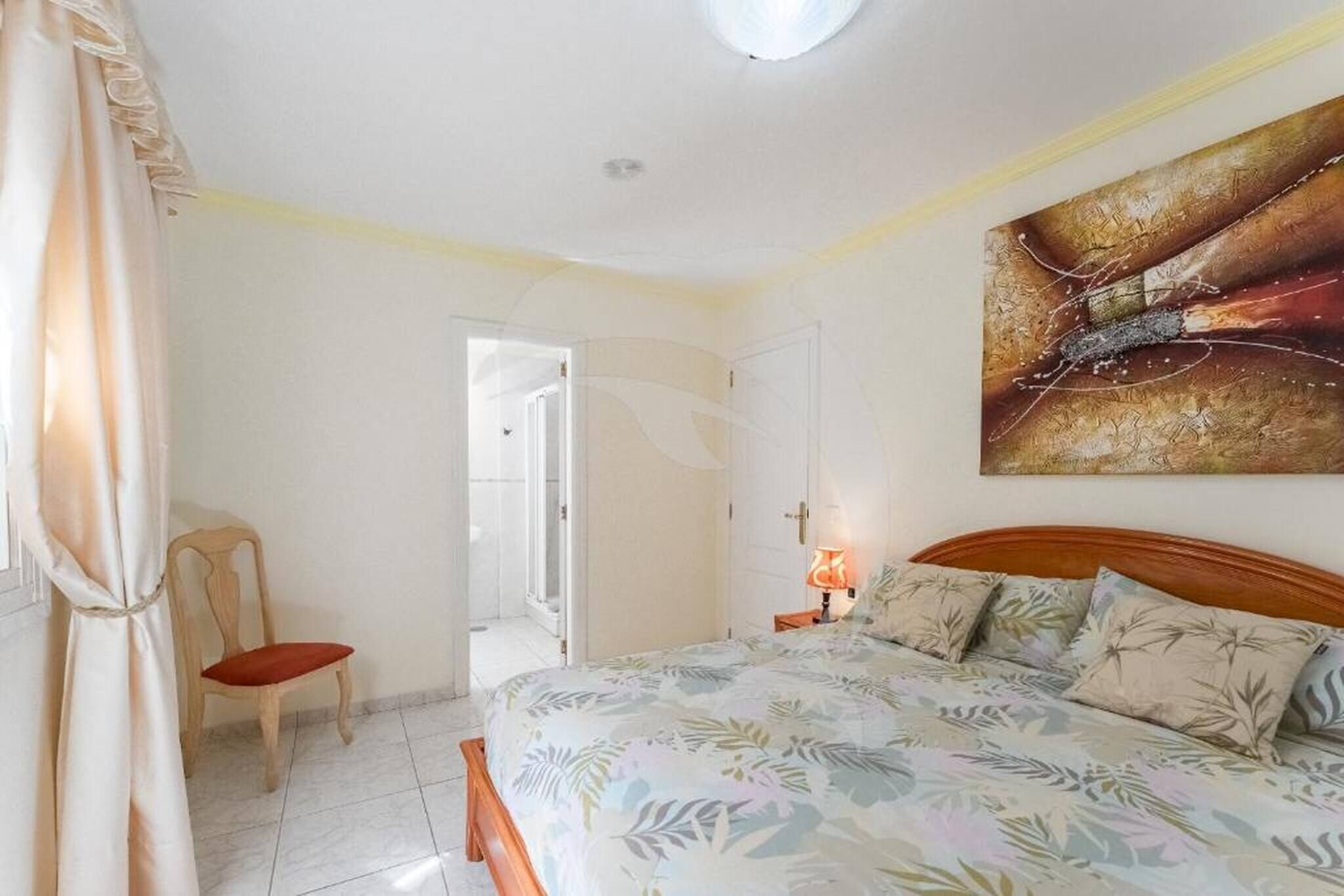 A bedroom with a bed, a chair, a painting, and a door leading to another room.