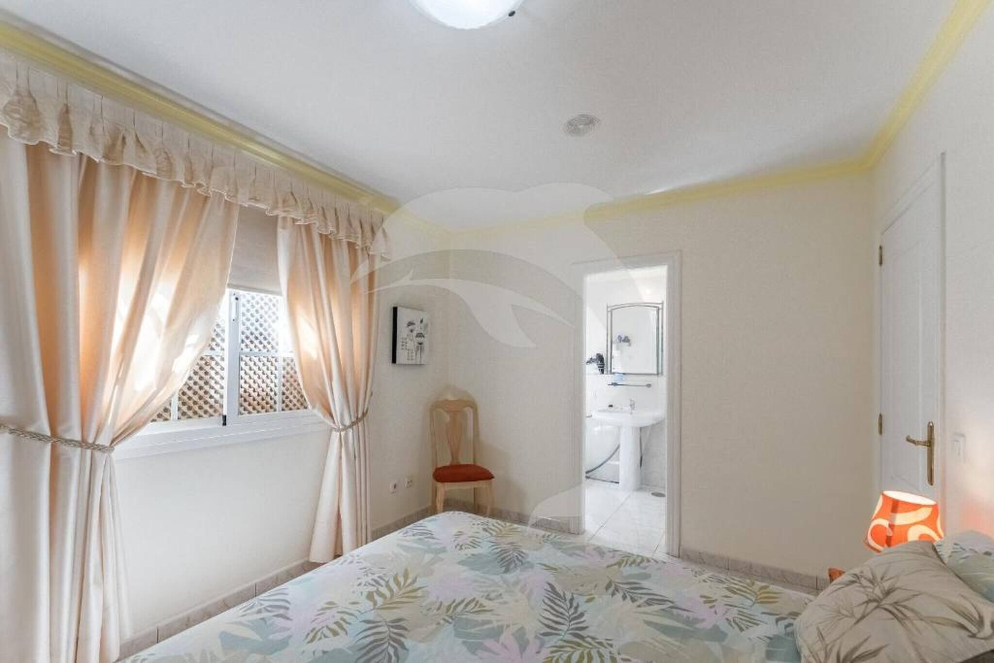 A bedroom with a bed, curtains, a chair, a lamp, and a bathroom visible through an open door.