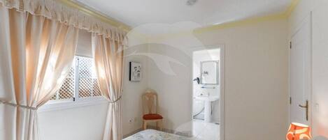 Un dormitorio con cama, cortinas, una silla, una lámpara y un baño visible a través de una puerta abierta.