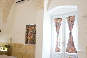 Ein Schlafzimmer mit einem Bett, einem Nachttisch mit Lampe, einem Fenster mit floralem Vorhang und einem Wandbild.