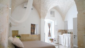 A bedroom with a bed, a small table, and a view of the kitchen.
