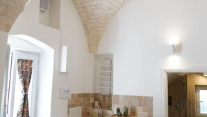 A bathroom with a shower, a sink, and a small shelf with toiletries. There is a towel rack and a window with a curtain.