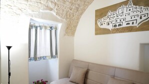 A room with a beige sofa, a window with curtains, a wall-mounted artwork of a building, and a small plant on the windowsill.