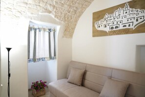 Une pièce avec un canapé beige, une fenêtre avec des rideaux, une œuvre d’art représentant un bâtiment accrochée au mur, et une petite plante posée sur le rebord de la fenêtre.