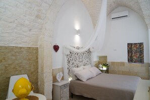 Une chambre avec un mur en pierre, un lit à baldaquin blanc, une table de chevet ornée d’un petit objet en forme de cœur et un climatiseur fixé au mur.