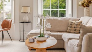 Een woonkamer met een beige bank, een ronde houten salontafel en een witte orchidee. Er staat een lamp op een bijzettafeltje, een stoel en een raam met uitzicht op groen.