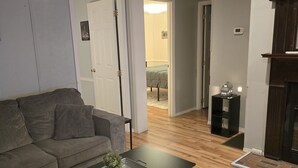 A modern living room with a grey sofa, a black coffee table with a tray and plant, a patterned rug, wooden flooring, and a visible hallway leading to another room.