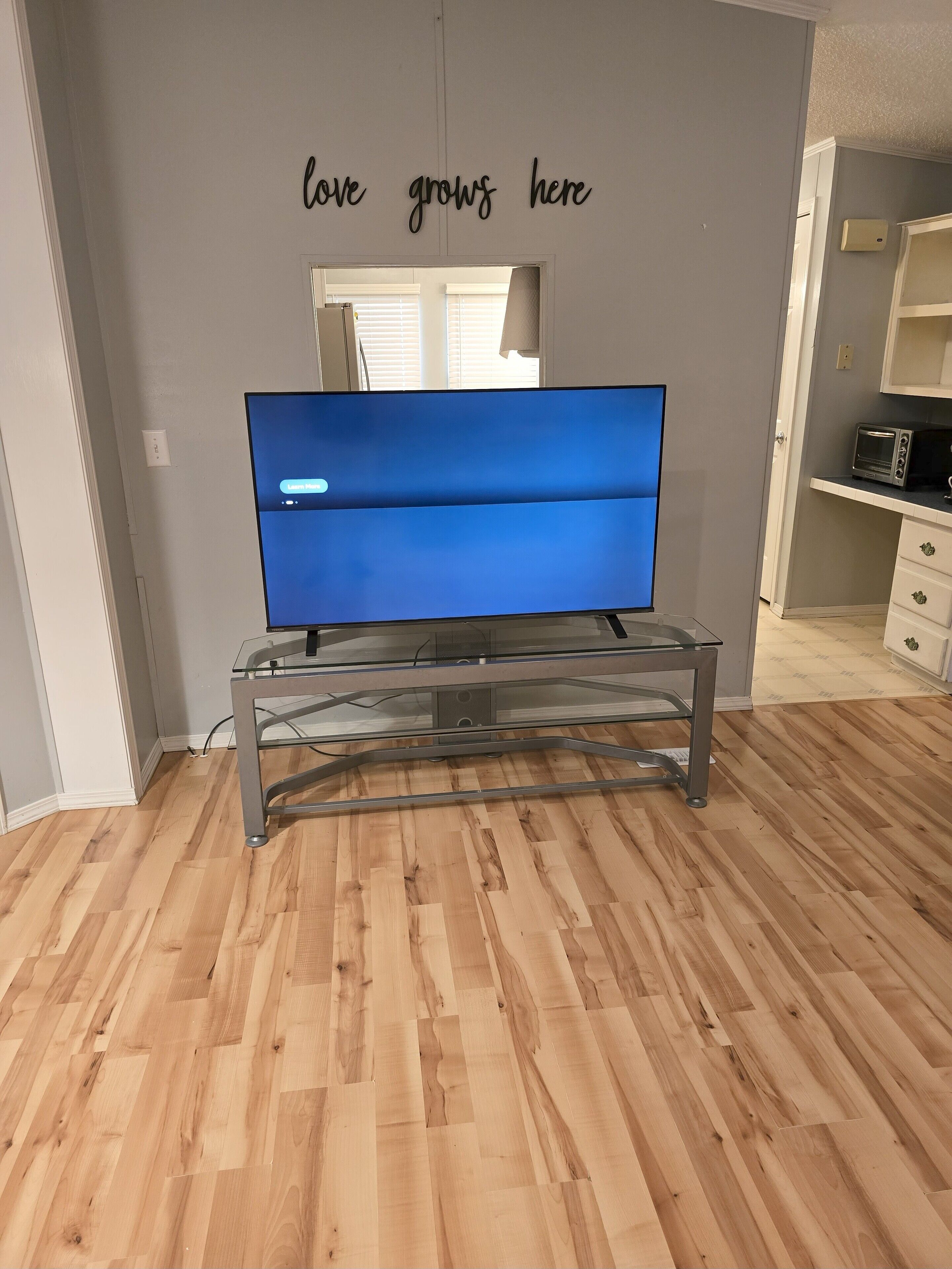Een kamer met een flatscreen televisie op een glazen tafel, een muur met de tekst 'love grows here' en een houten vloer.