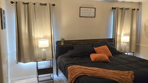 A bedroom with a black bed, two bedside lamps, a bedside table with a plant, and a wall-mounted sign.