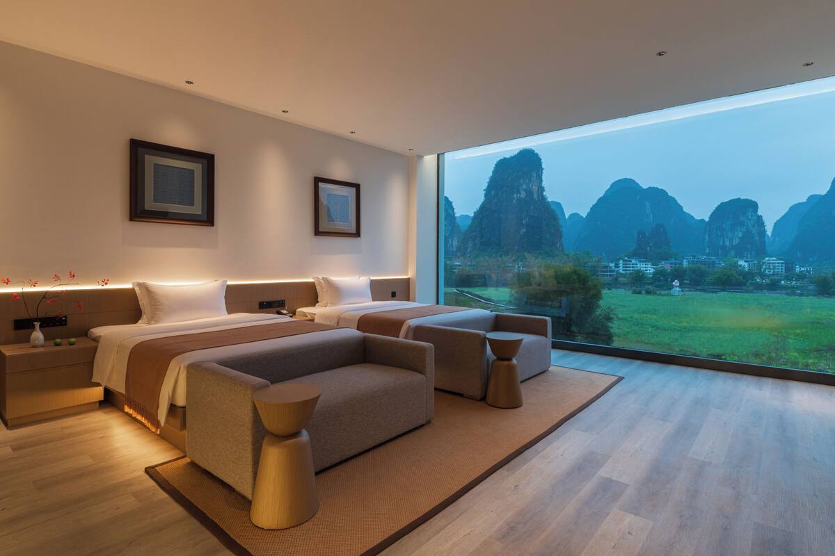 A modern hotel room with a large bed, two bedside tables, a sofa, and a view of a mountainous landscape.